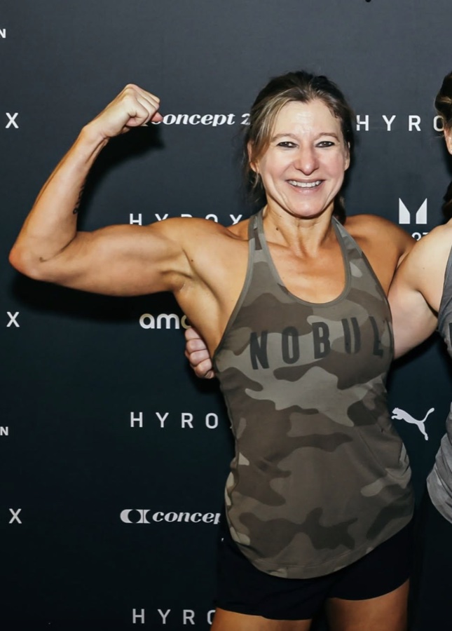Unique+Fit trainer flexing in the studio, surrounded by kettlebells, sandbags, and battle ropes